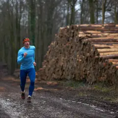 Mężczyzna w niebieskiej odzieży biegający po leśnej ścieżce obok stosu drewna.