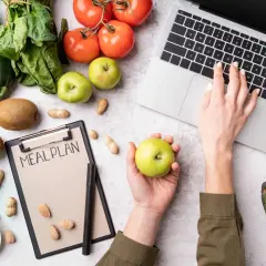 Dłoń z jabłkiem nad notatnikiem "MEAL PLAN", obok laptop, warzywa i owoce.
