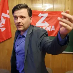 Mężczyzna w garniturze gestykuluje na tle czerwonych flag z białym symbolem "Z".