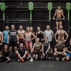 Grupa osób w strojach sportowych pozuje przed czarną ścianą w siłowni CrossFit.