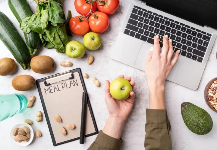Dłoń z jabłkiem nad notatnikiem "MEAL PLAN", obok laptop, warzywa i owoce.