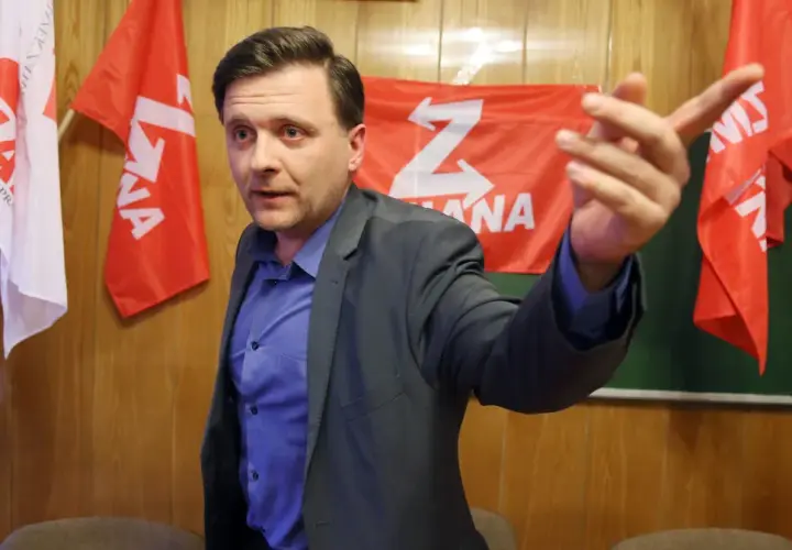 Mężczyzna w garniturze gestykuluje na tle czerwonych flag z białym symbolem "Z".