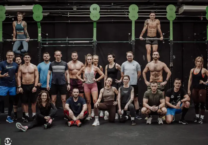 Grupa osób w strojach sportowych pozuje przed czarną ścianą w siłowni CrossFit.