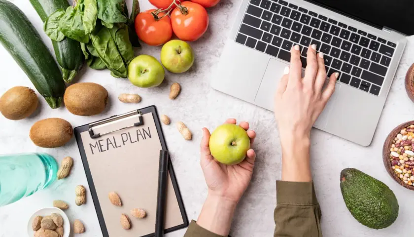 Dłoń z jabłkiem nad notatnikiem "MEAL PLAN", obok laptop, warzywa i owoce.