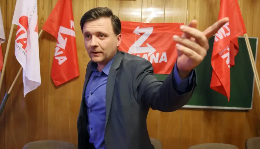 Mężczyzna w garniturze gestykuluje na tle czerwonych flag z białym symbolem "Z".