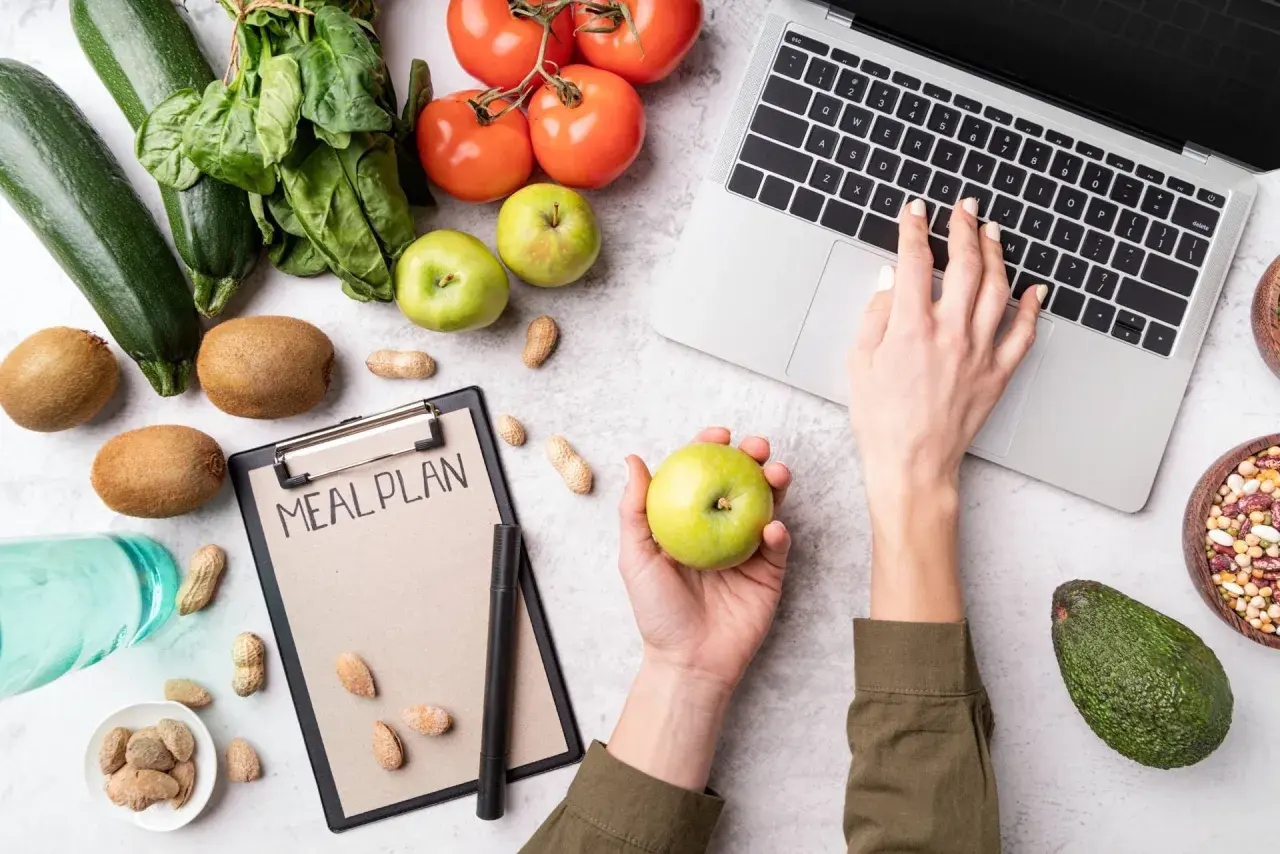Dłoń z jabłkiem nad notatnikiem "MEAL PLAN", obok laptop, warzywa i owoce.
