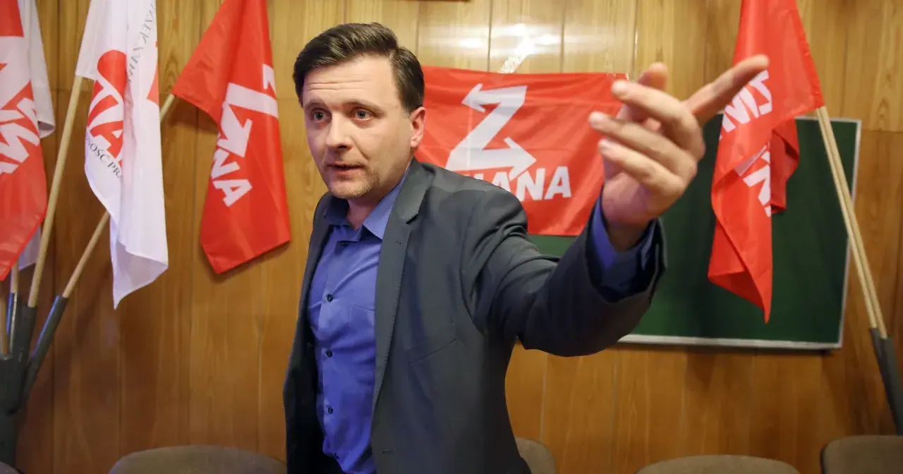 Mężczyzna w garniturze gestykuluje na tle czerwonych flag z białym symbolem "Z".