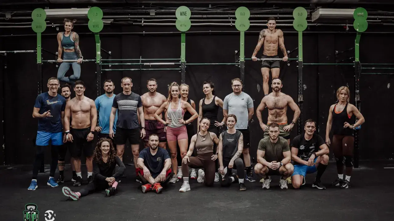Grupa osób w strojach sportowych pozuje przed czarną ścianą w siłowni CrossFit.